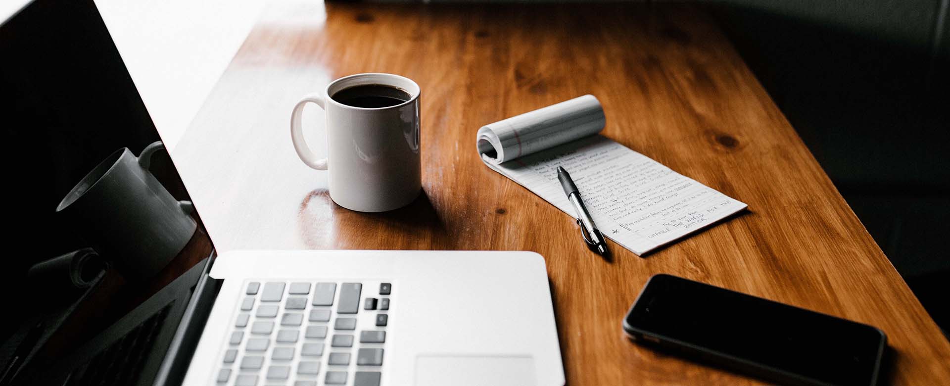 Laptop, coffee cup, notepad and phone on a wooden desk