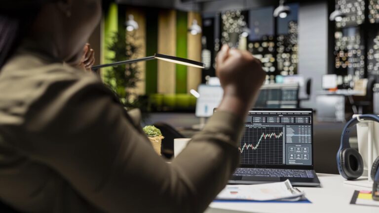 Person at a desk with a hand raised in celebration in front of a screen of charts