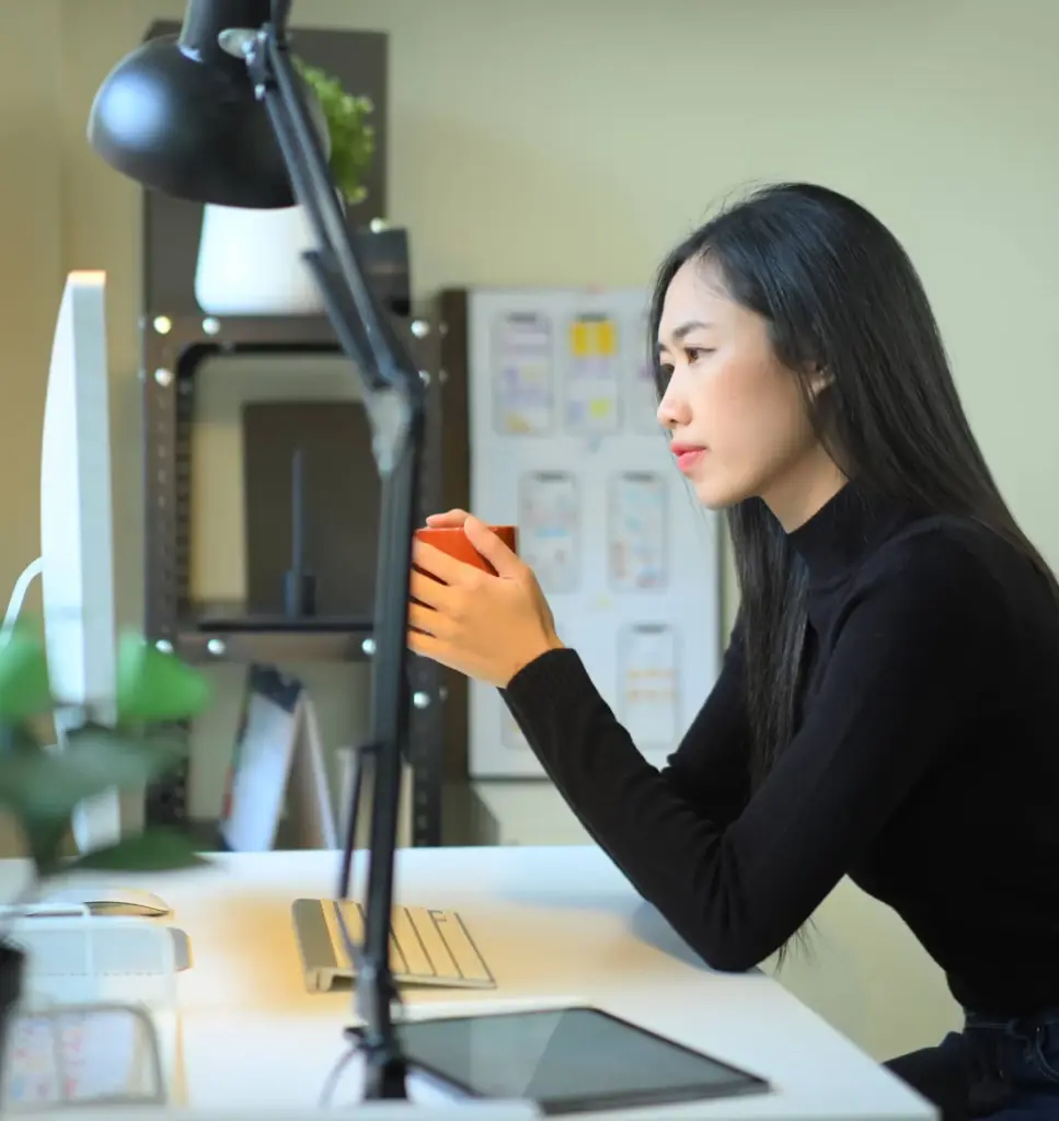 A strategist at her desk reviewing work on screen, with page wireframes pinned to the wall behind her