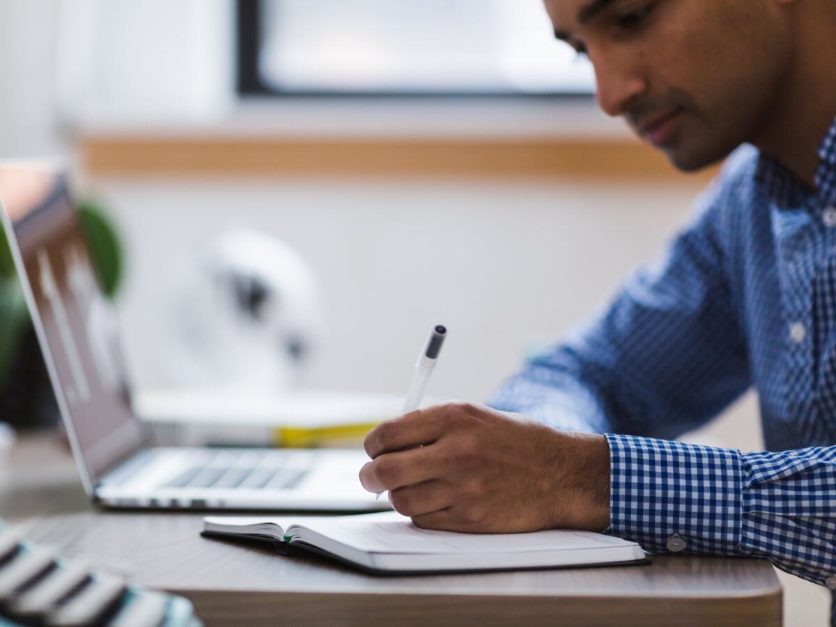Man writing notes in a notebook beside his laptop