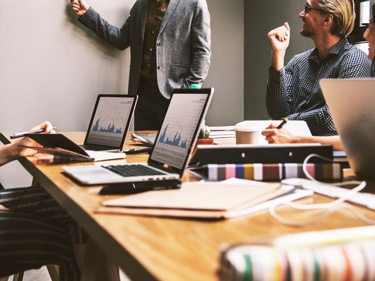 Team around a meeting table with laptops while a colleague points at the wall