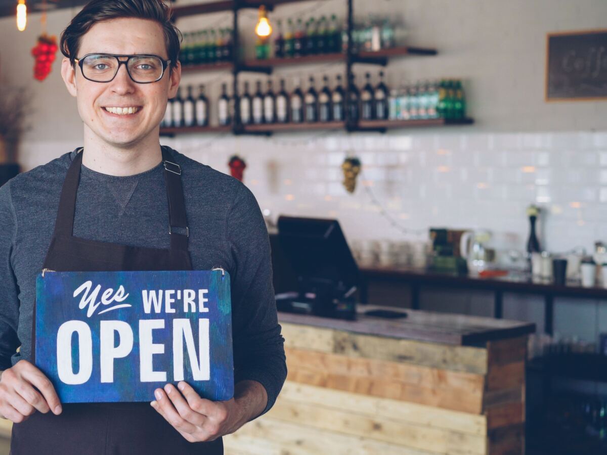 Smiling shop owner holding a Yes We are Open sign behind the counter