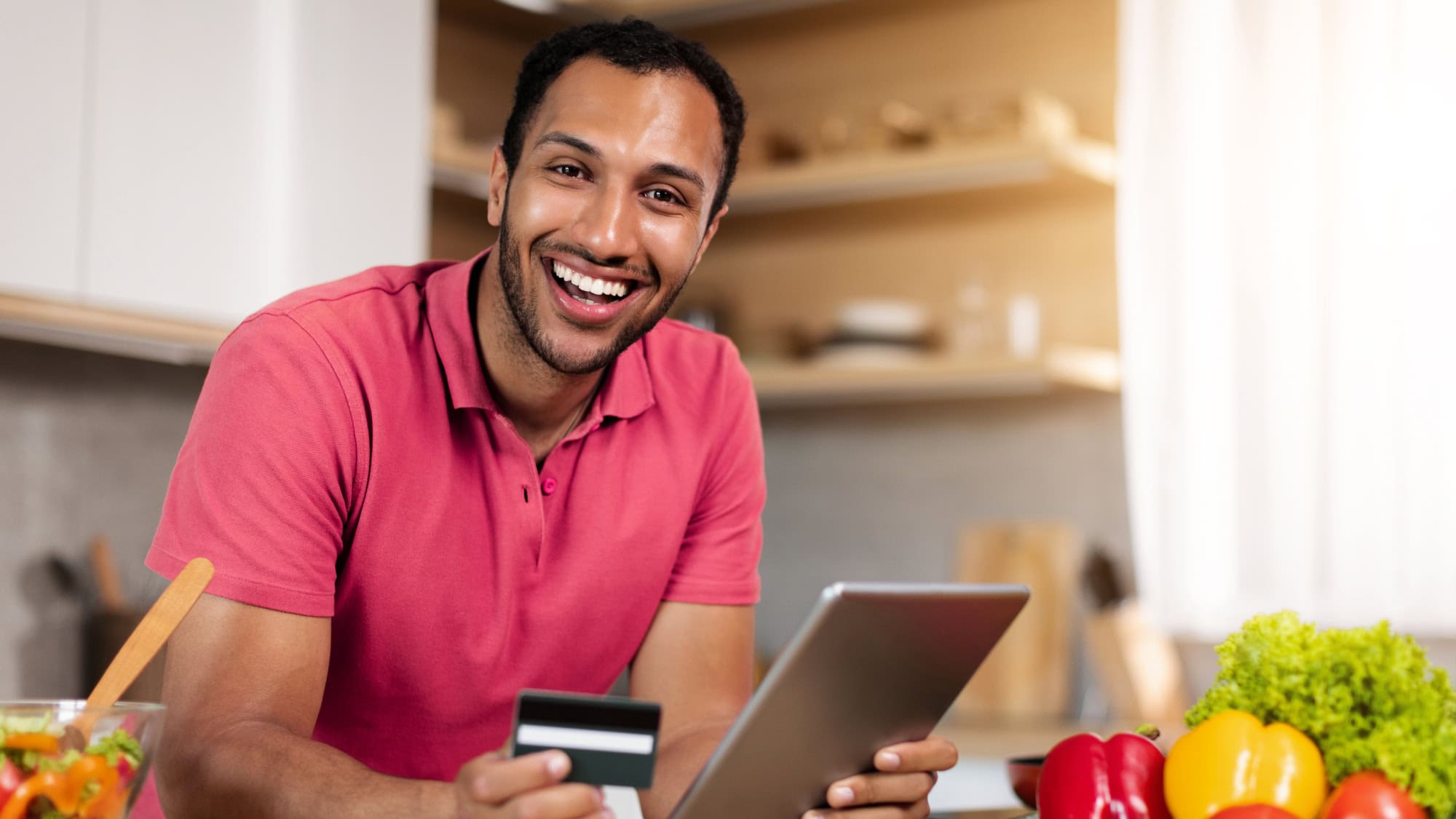Smiling man in his kitchen paying online with a card and a tablet