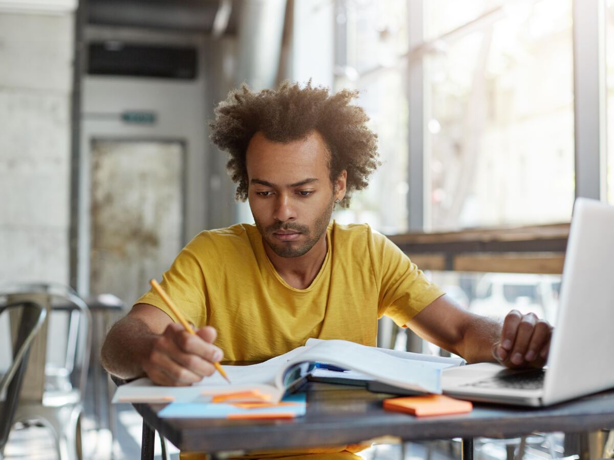 Young man in a yellow shirt working with a notebook and laptop in a cafe